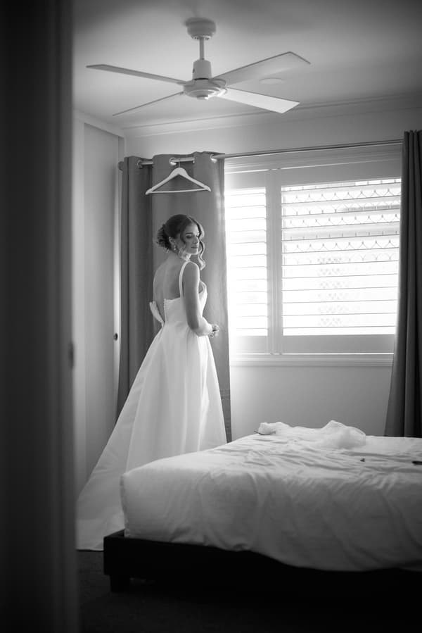 The bride Ashleigh stands near a window with curtains in a bedroom, wearing her wedding gown at Sandstone Point Hotel.