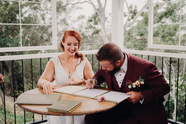 The bride and groom sign their wedding documents at a round wooden table on the ceremony stage at Sandstone Point Hotel — The Pavilion, with greenery visible outside the railing.