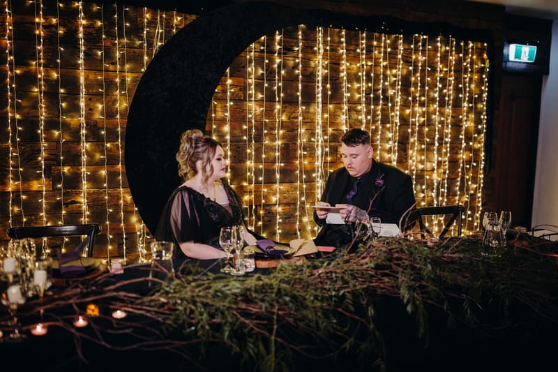 The bride Renae and groom Adam sit at a decorated table with greenery and candles in the Ocean View Estates — Function Room, with a backdrop of string lights and a large black crescent moon decoration.