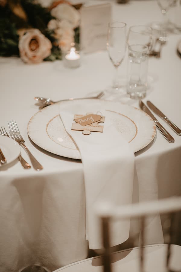 Close-up of a wedding reception place setting with a white plate, white napkin, cutlery, glassware, a small name tag reading 'Penoy', and a lit candle in the background.