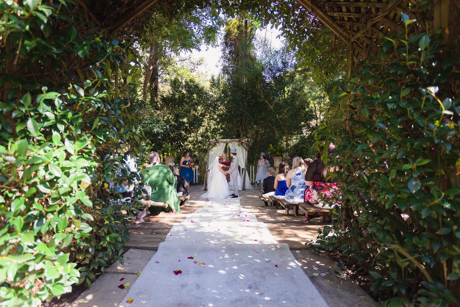 Bride Holly and groom Paul stand at the altar exchanging vows at Kwila Lodge, surrounded by seated guests on wooden benches under a leafy archway.