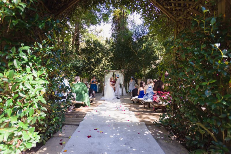 Bride Holly and groom Paul stand at the altar exchanging vows at Kwila Lodge, surrounded by seated guests on wooden benches under a leafy archway.