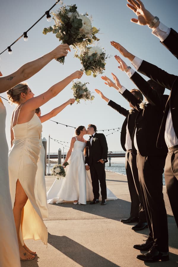 Bride Ashleigh and groom James kiss while holding hands at Sandstone Point Hotel, framed by bridesmaids raising bouquets and groomsmen raising arms in celebration near the waterfront.