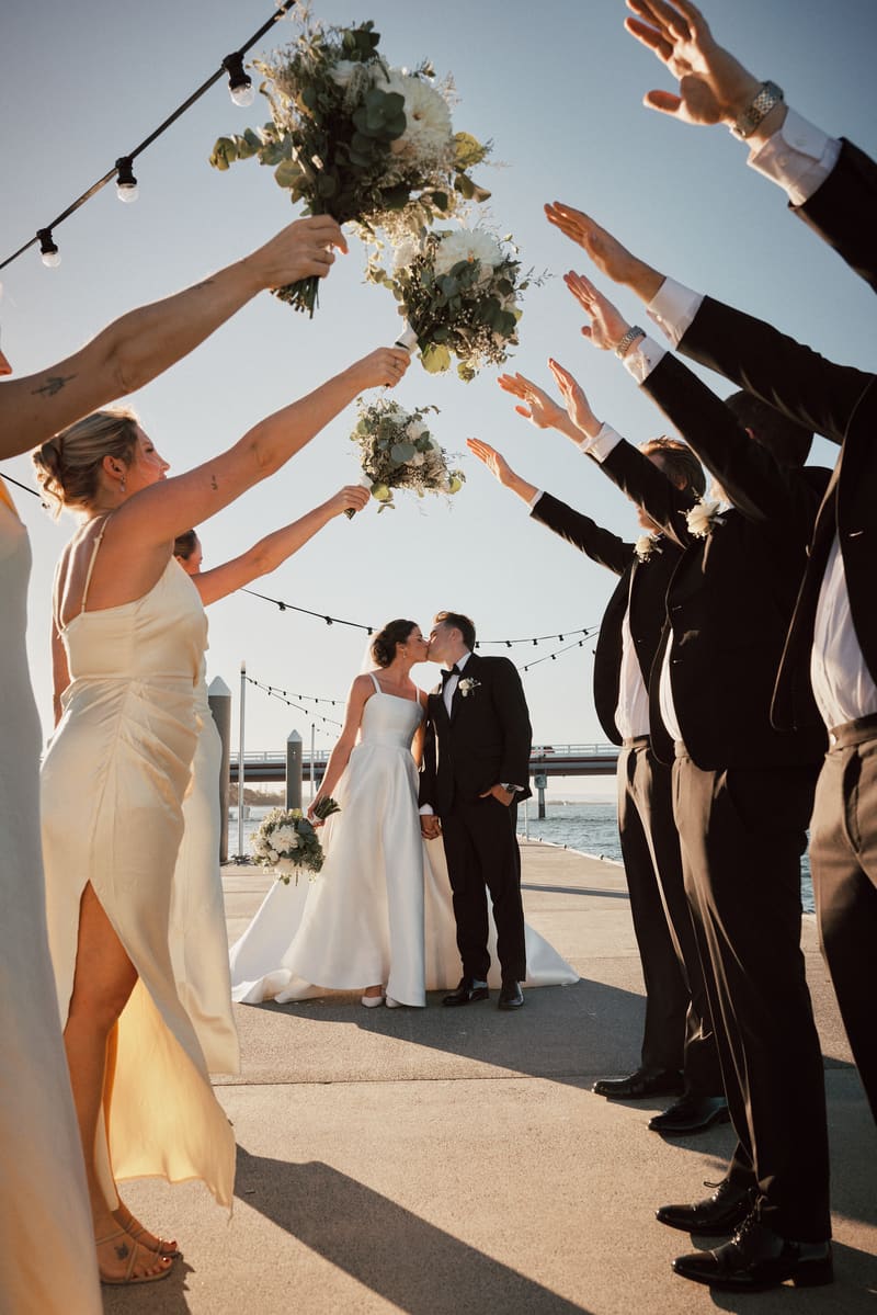 Bride Ashleigh and groom James kiss while holding hands, framed by bridesmaids raising bouquets and groomsmen raising arms in celebration near the waterfront. Best Brisbane wedding photographer by She Said Yes.