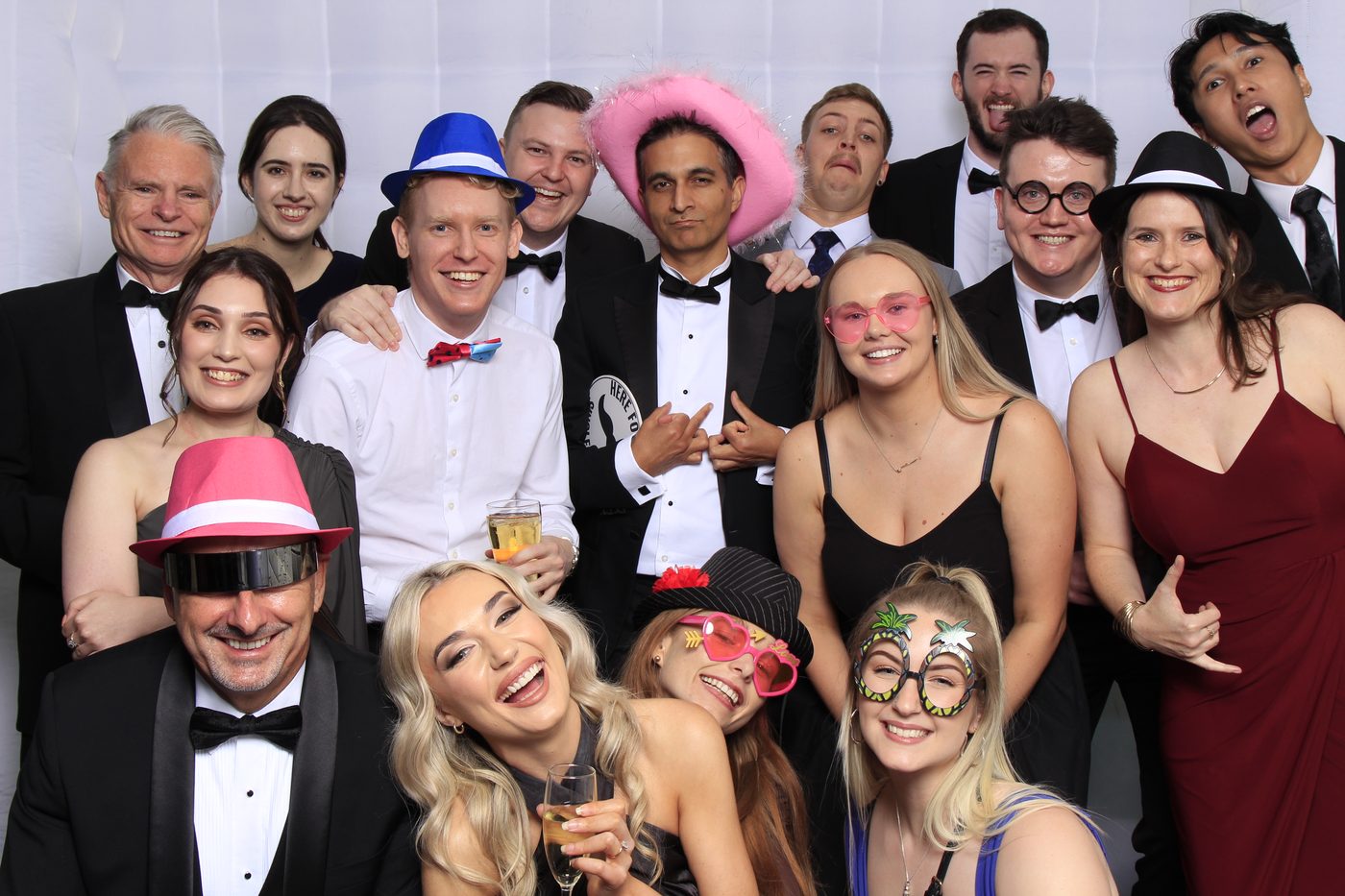 Group of guests in formal wear with fun props and hats, smiling in a photo booth.