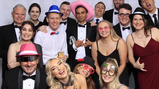 Group of guests in formal wear with fun props and hats, smiling in a photo booth.