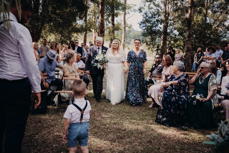 The bride Annie walks down the aisle at Ocean View Estates, accompanied by an older man and woman, with guests seated on either side watching the procession.