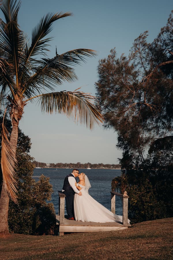 The bride Chantelle and groom Peter share a kiss on a small wooden platform overlooking the water at Sandstone Point Hotel, framed by trees and a palm tree.
