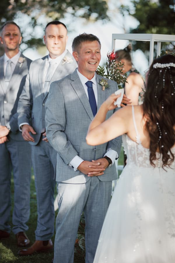 Dane, the groom, stands smiling facing Jheng, the bride, who is wiping her eyes during the ceremony at Sandstone Point Hotel — The Pavilion. Two groomsmen stand behind Dane.