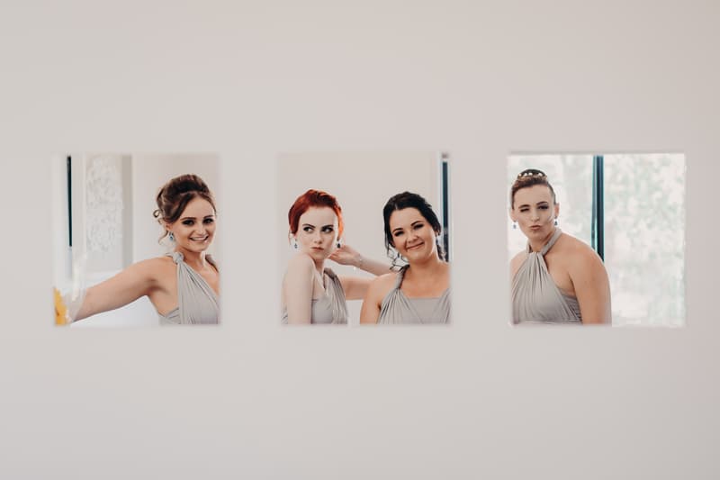 Three bridesmaids pose individually behind three square cutouts in a white wall at Ocean View Estates.