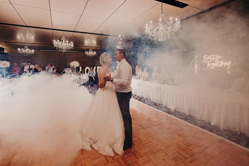 Emily and Dylan share their first dance surrounded by fog on the dance floor at the Sandstone Point Hotel — Pumicestone Room, with guests seated at tables in the background and a 'Better Together' neon sign visible.