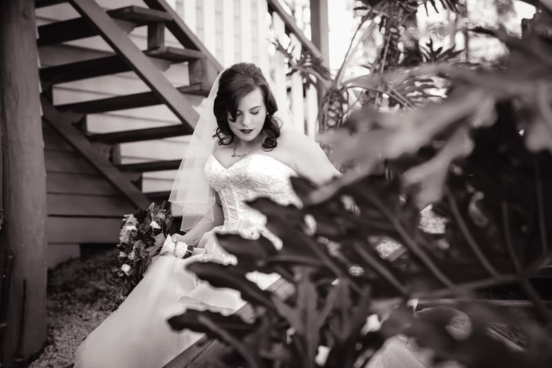 The bride Holly sits on a bench near a staircase at Kwila Lodge, holding her bouquet and looking down.