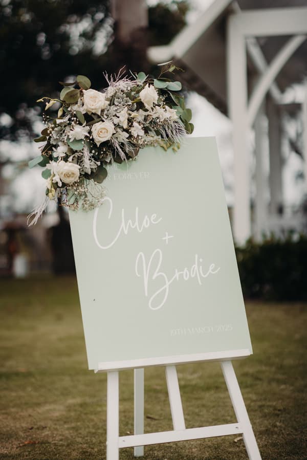 Wedding welcome sign for Chloe and Brodie at Sandstone Point Hotel, decorated with white roses and greenery on a white easel outdoors.