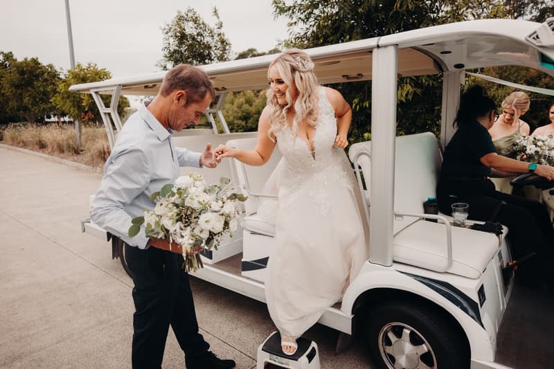 The bride Chloe steps down from a white golf cart with a man holding her bouquet and offering his hand for support at Sandstone Point Hotel — Pavilion. Two bridesmaids sit inside the golf cart.