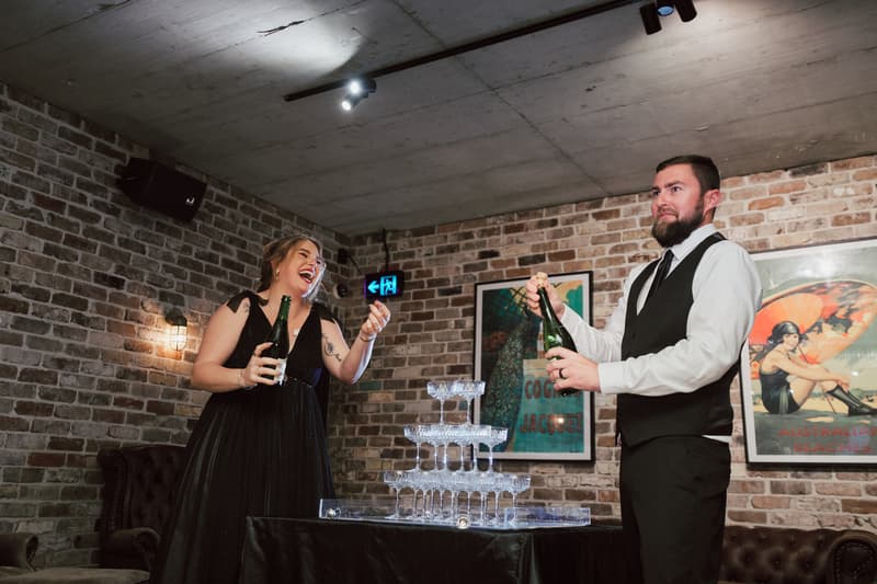 The bride and groom at Sandstone Point Hotel — The Cellar prepare to pour champagne into a stacked pyramid of glasses during the reception.