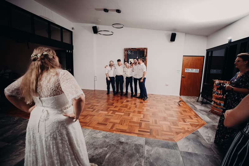 The bride Annie stands with hands on hips facing five men in white shirts and dark pants posing together on the dance floor at Ocean View Estates reception stage, while two female guests watch from the right side.