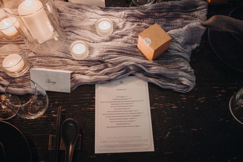 Table setting at Donna Chang with a banquet menu, candles, a place card labeled 'Terry', a small gift box, and glassware on a dark wooden table with a textured cloth runner. Full day wedding photography by She Said Yes.