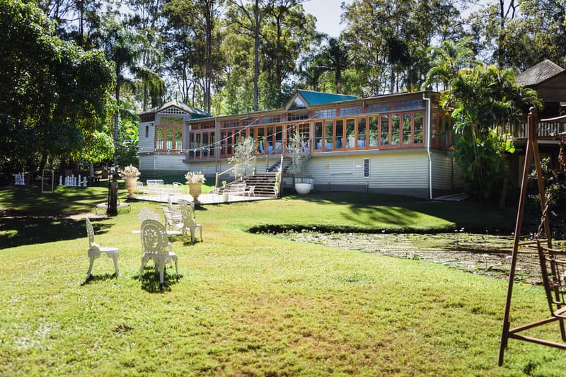 Exterior view of Kwila Lodge showing a grassy lawn with white chairs arranged and a pond in the foreground, surrounded by trees and greenery.
