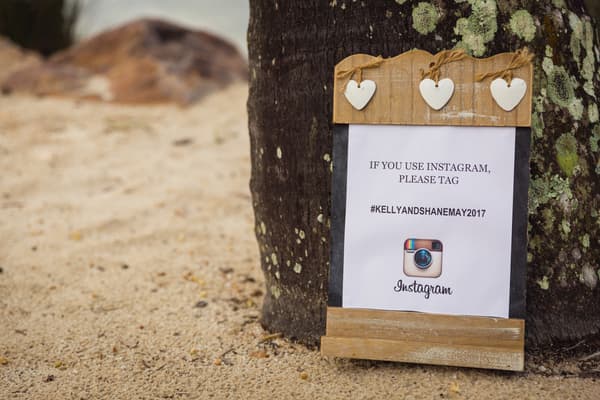 A wooden sign with three white heart decorations is placed against a tree trunk at Novotel Sunshine Coast Resort, displaying a printed note asking guests to tag Instagram posts with #KELLYANDSHANEMAY2017.