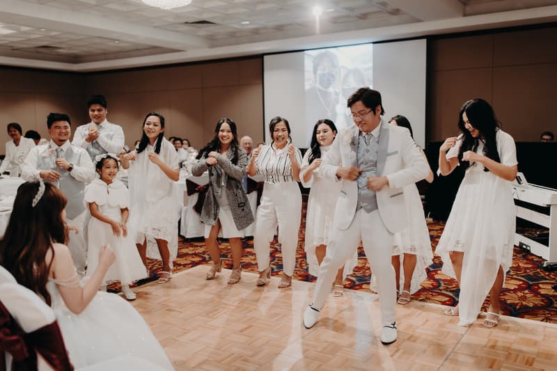 The groom Timothy dances on the dance floor at the Royal on the Park reception, surrounded by guests and bridesmaids in white dresses, while the bride Henny watches seated in the foreground.