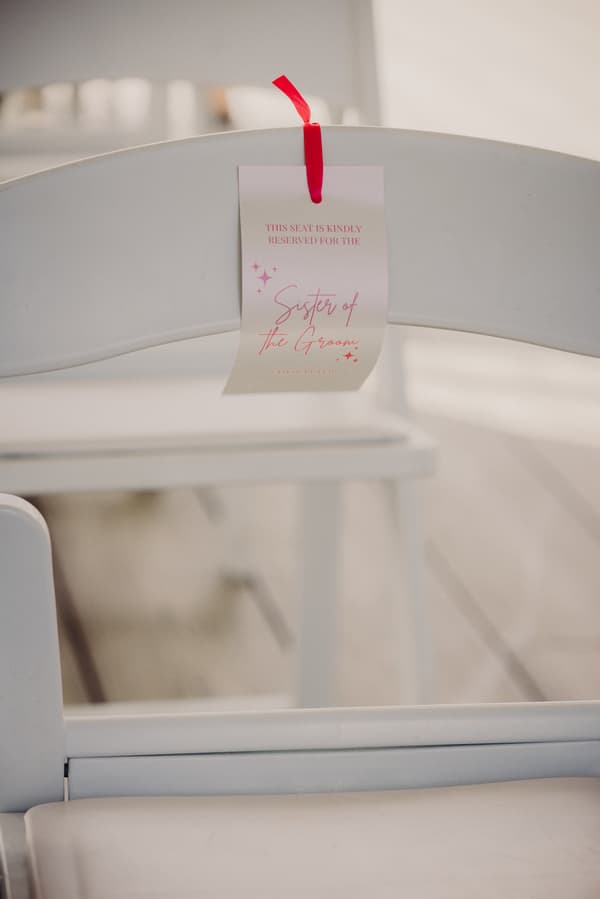 A white folding chair at Sandstone Point Hotel — Pavilion with a tag attached by a red ribbon reading 'This seat is kindly reserved for the Sister of the Groom.'