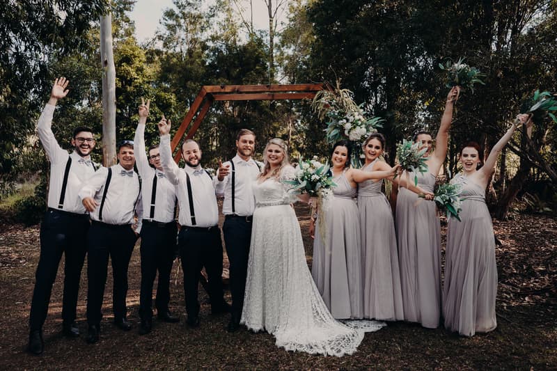 Annie and Trent pose with their bridal party at Ocean View Estates, with the bridesmaids in light gray dresses holding bouquets and the groomsmen in white shirts with suspenders and bow ties, standing outdoors in front of a wooden arch.