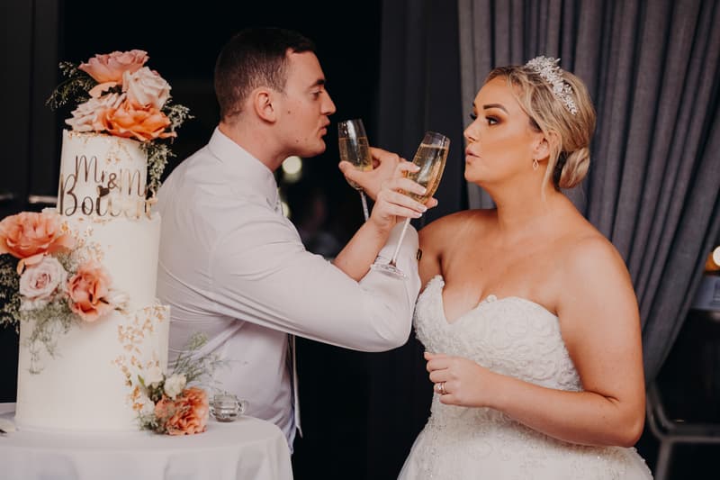 Bride Emily and groom Dylan toast with champagne glasses next to a three-tier wedding cake decorated with peach and pink flowers at Sandstone Point Hotel — Pumicestone Room.