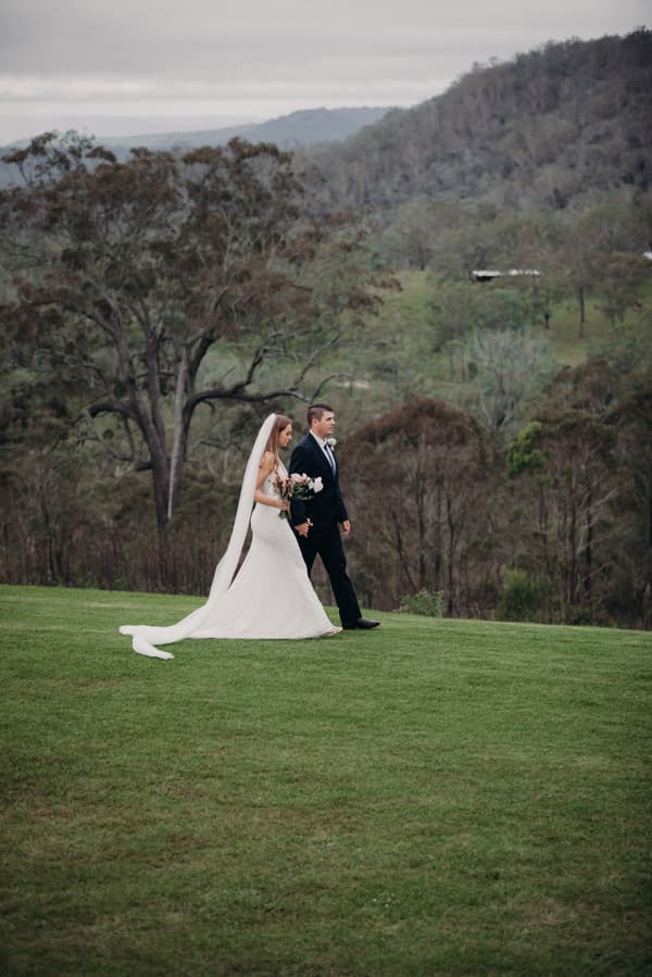 Chelsea in a white wedding gown holding a bouquet and Brent in a black suit walk hand in hand on a grassy hill at Preston Peak Winery Functions Centre with trees and hills in the background.