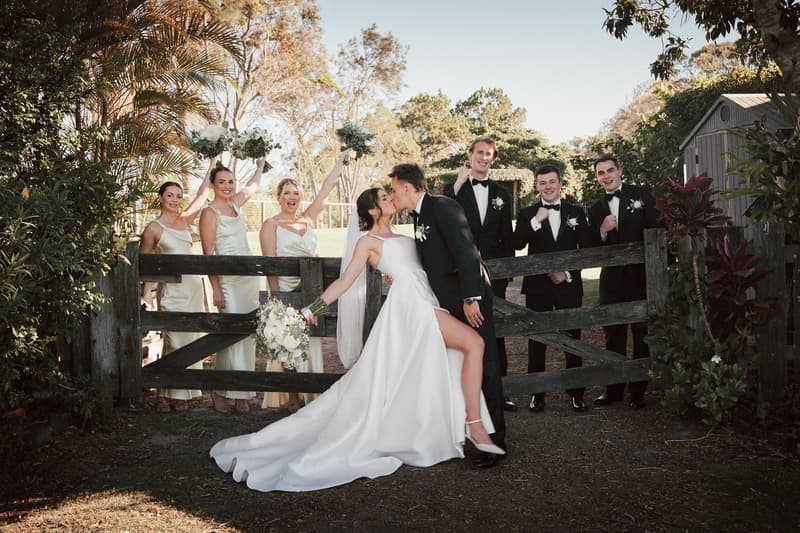 Bride Ashleigh and groom James kiss in front of a wooden gate at Sandstone Point Hotel, with three bridesmaids holding bouquets on the left and three groomsmen in tuxedos on the right.