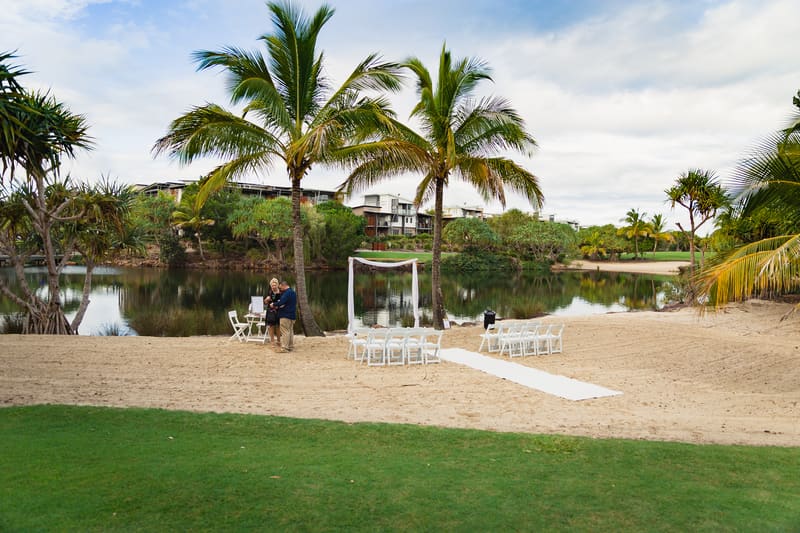 Wedding ceremony setup on a sandy area, a white aisle runner, and two people standing near a small table by the water. Brisbane videography by She Said Yes.