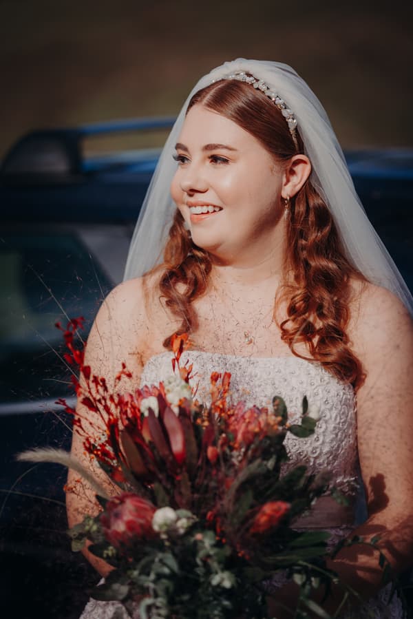 The bride Lilly in a white strapless wedding dress and veil holding a bouquet of red and green flowers at Yabbaloumba Retreat — By The River.