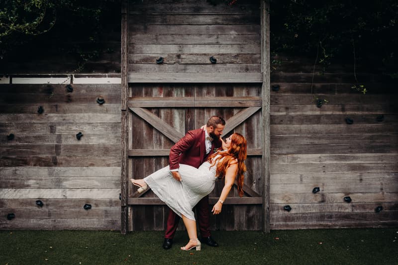 Jayden dips the bride Red in front of a large rustic wooden door at Sandstone Point Hotel — The Pavilion.