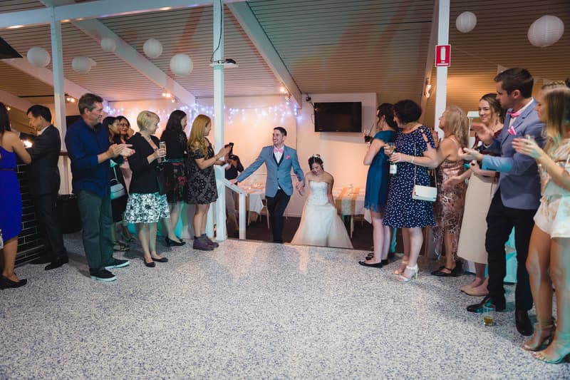 Bride Wing and groom Jason enter the reception stage at Bilinga Beach Weddings — Bilinga SLSC, greeted by applauding guests on either side.