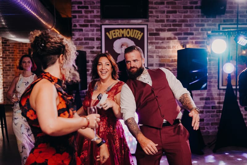 The bride in a red dress and the groom in a burgundy vest and pants dance together at the reception stage of Sandstone Point Hotel — The Cellar, with guests visible in the background.