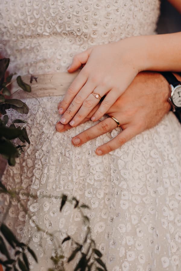 Close-up of the bride's and groom's hands resting on the bride Lilly's beaded wedding dress, showing their wedding rings.