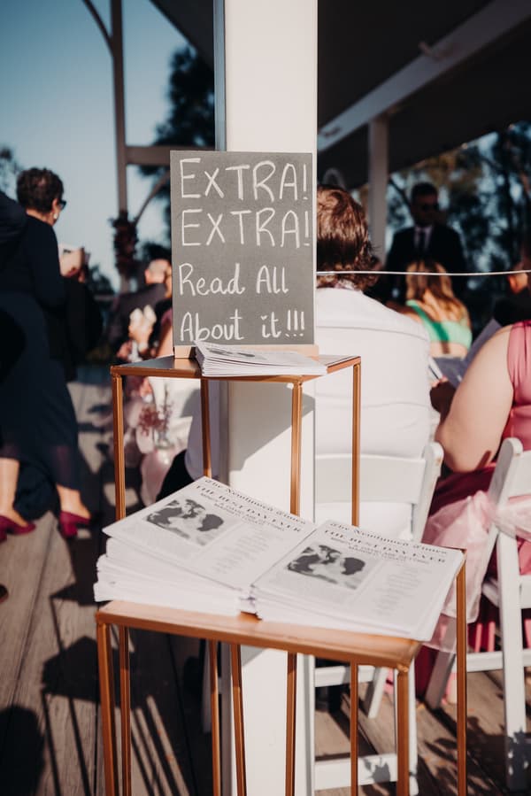 A stack of wedding newspapers titled 'The Wedding Gazette' is displayed on a gold stand with a sign reading 'EXTRA! EXTRA! Read All About it!!!' at Sandstone Point Hotel — Pavilion. Guests are seated on white chairs in the background.