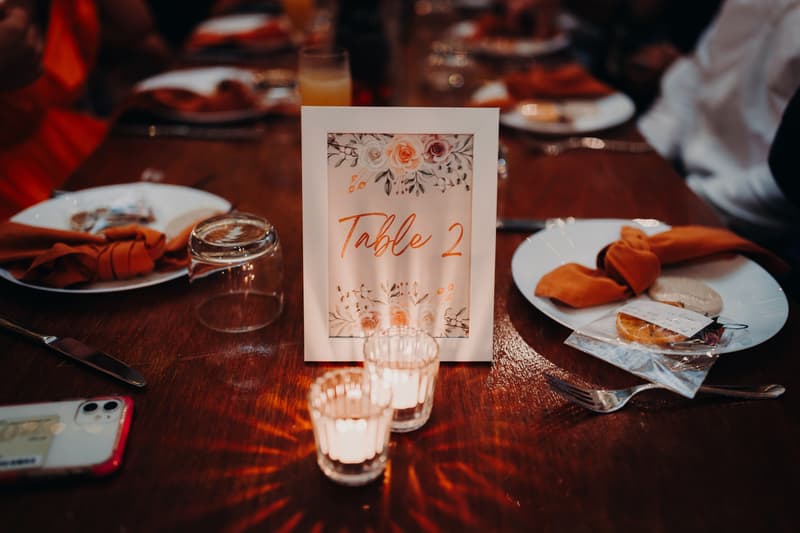 Table 2 at the reception stage of Lilly and Connor's wedding at Yabbaloumba Retreat — The Shed, featuring a floral table number card, two lit votive candles, place settings with orange napkins, and a smartphone on the wooden table.