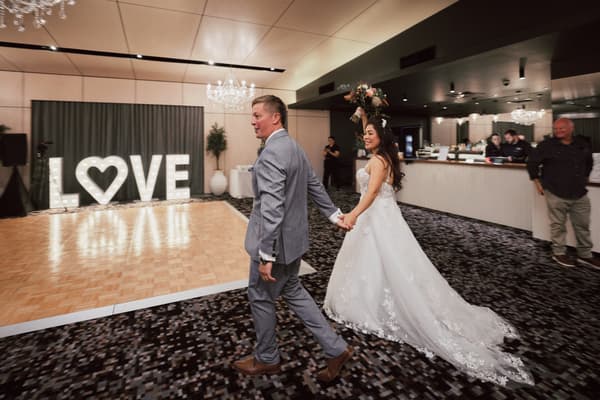 Bride Jheng and groom Dane hold hands walking near the dance floor, with illuminated LOVE letters in the background and guests near the bar. Winter wedding light photography Brisbane by She Said Yes.