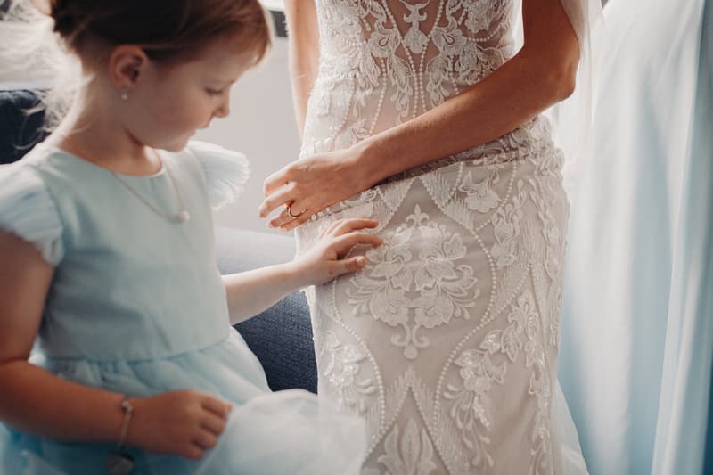 The bride Courtney is shown from the waist down wearing a detailed lace wedding dress, with a young flower girl in a light blue dress touching the fabric.