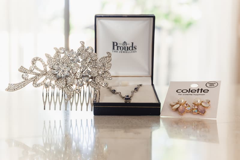 Close-up of bridal jewelry including a rhinestone hair comb, a necklace in a Prouds The Jewellers box, and a pair of Colette earrings displayed on a marble surface at Kwila Lodge.