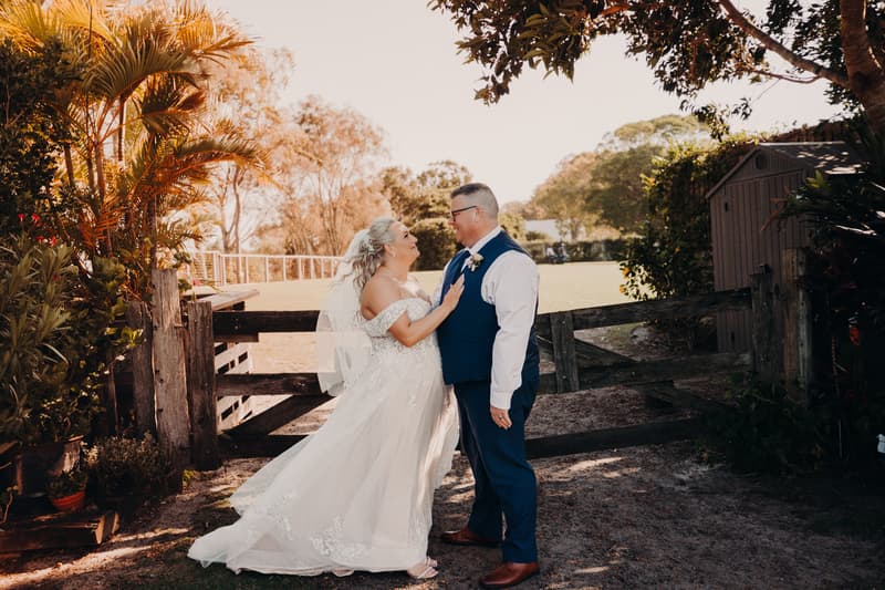 The bride Chantelle and groom Peter stand facing each other near a wooden gate surrounded by greenery at Sandstone Point Hotel during their couple portraits session.