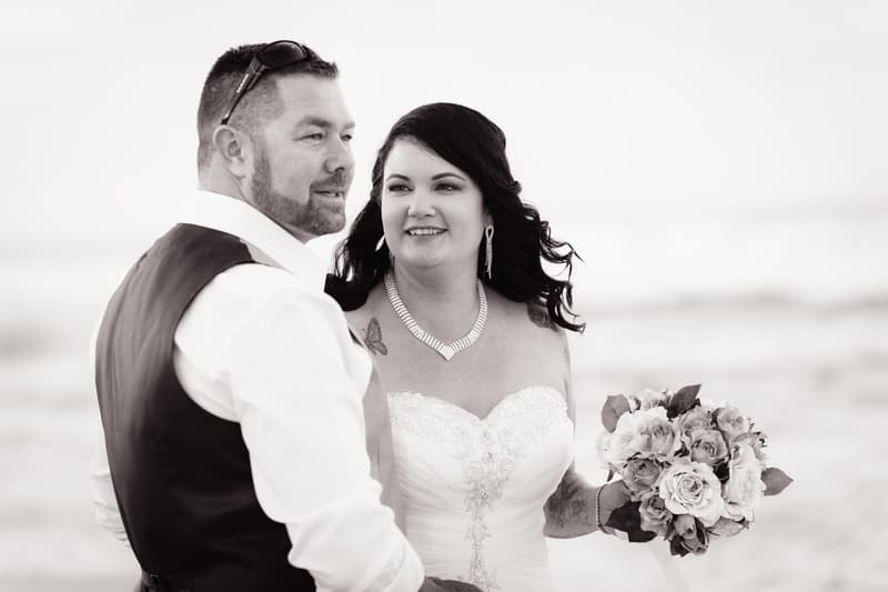 Bride Kelly in a strapless wedding gown holding a bouquet stands next to groom Shane wearing a white shirt and dark vest, both looking slightly off-camera in an outdoor setting.