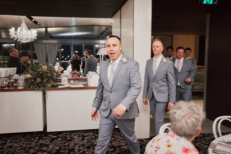 The groom and groomsmen walk into the reception room at Sandstone Point Hotel — The Pumicestone Room, dressed in matching grey suits with boutonnieres.