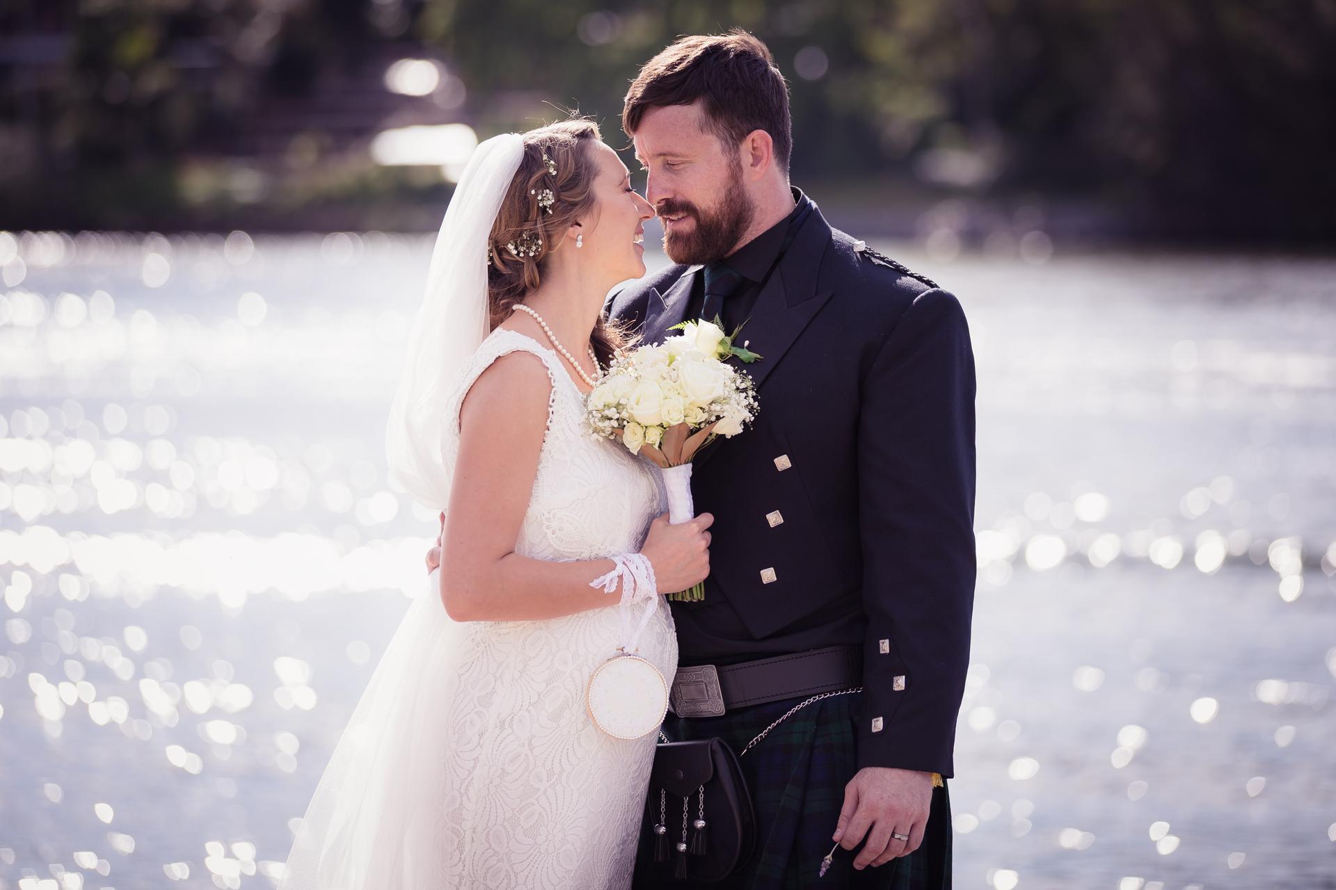 Bride Francesca and groom Ben embrace near a body of water at Toowong Rowing Club, The Malouf Room, with Francesca holding a bouquet of white flowers and Ben wearing a traditional kilt outfit.