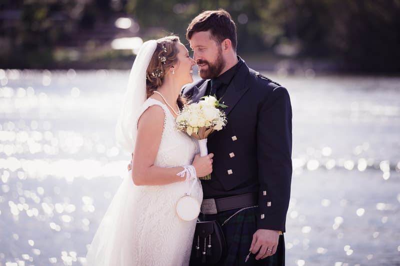 Bride Francesca and groom Ben embrace near a body of water at Toowong Rowing Club, The Malouf Room, with Francesca holding a bouquet of white flowers and Ben wearing a traditional kilt outfit.