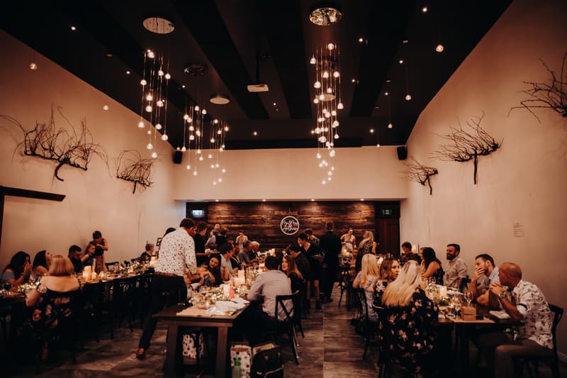Guests are seated at long tables enjoying the wedding reception at Ocean View Estates, with warm hanging lights and wall decorations visible.