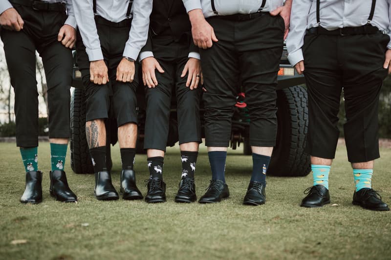 Five groomsmen stand side by side on grass with their pant legs rolled up, showing colorful patterned socks and black shoes, in front of a vehicle at Sandstone Point Hotel — The Pavilion.