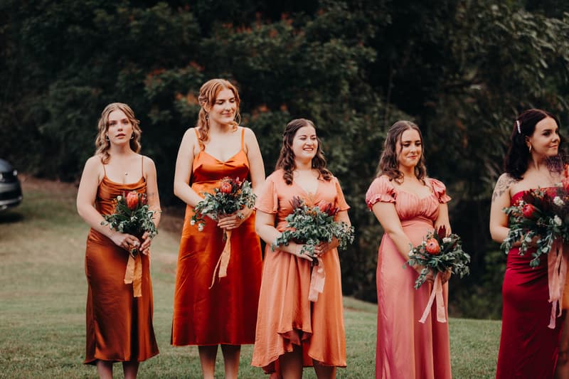Five bridesmaids stand outdoors holding bouquets at Yabbaloumba Retreat — By The River, dressed in various shades of orange, pink, and red gowns.