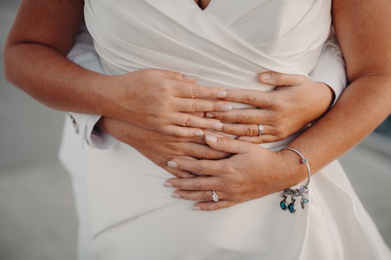 Brooke and Tiffany's hands intertwined over Brooke's white wedding dress, showing their wedding rings and a charm bracelet on Tiffany's wrist.