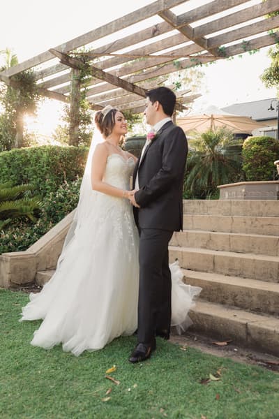 Bride Maryam and groom Pasha stand holding hands near stone steps under a wooden pergola, surrounded by greenery and soft sunlight. Elopement photography Brisbane by She Said Yes.